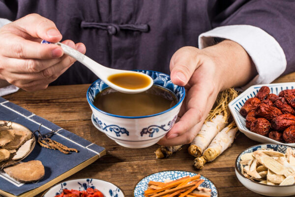 a man wearing vintage chinese clothing drinking a bowl of traditional chinese medicine herbal soup, chinese herbs surrounding the table